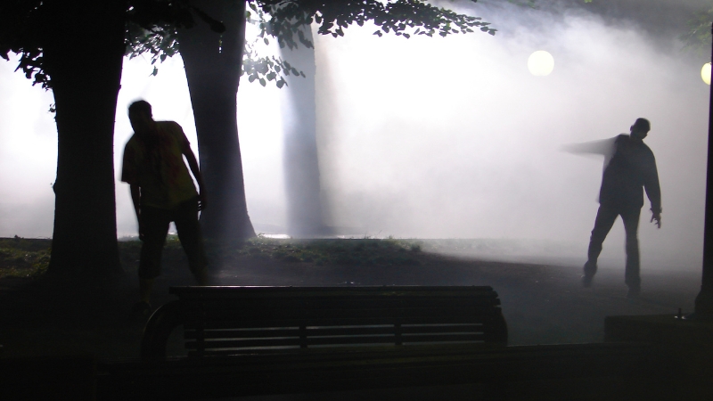 Dve siluete nalik zombijima kreću se kroz magloviti park noću, uz klupu i drveće u prvom planu